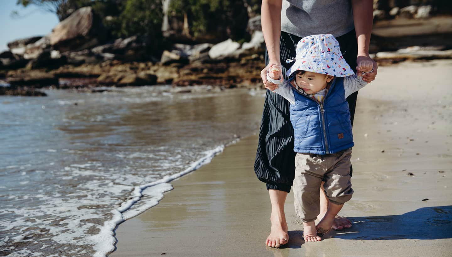 New Beginnings mother helping toddler walk along beach