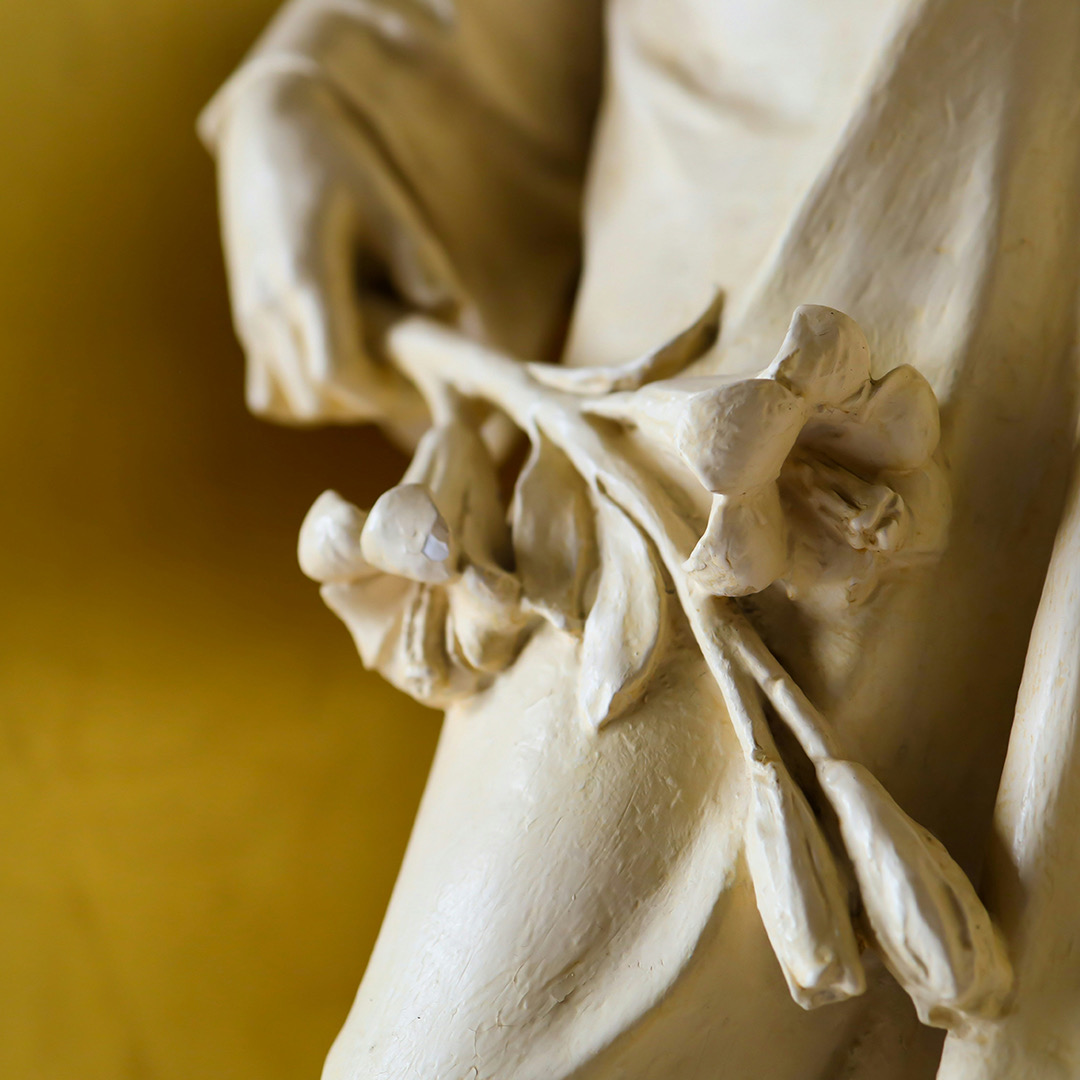 Part of a white stone statue. A hand holds three flowers at a downward angle across the body. The background is a faded yellow.