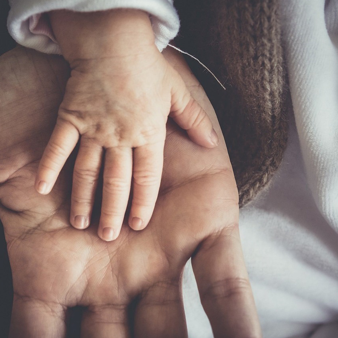 An infant's hand lays on the palm of a larger hand. Both hands lay on a white cloth surface.