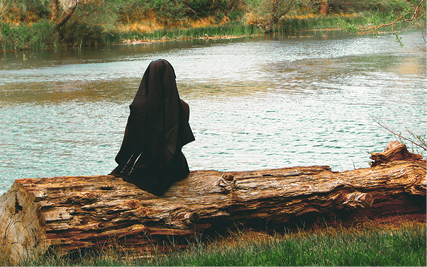 A Carmelite Sister sits on a fallen log facing away over a river.